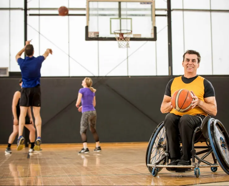 A basketball player in a wheelchair holds a basketball and smiles at the camera. Behind him are three other people playing basketball, with one person shooting a goal. They are all in an indoor basketball court.